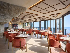 A restaurant with red velvet chairs, round tables, and a view of the sea.