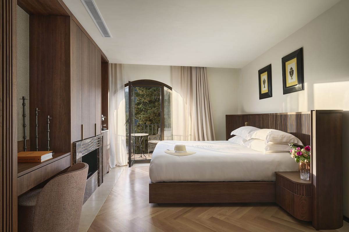 A spacious bedroom with a large bed, a wooden wardrobe, a small table with a vase, and a view of greenery through a window.