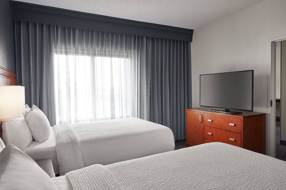 Une chambre d’hôtel avec deux lits, un téléviseur posé sur un meuble en bois et une grande fenêtre avec des rideaux.