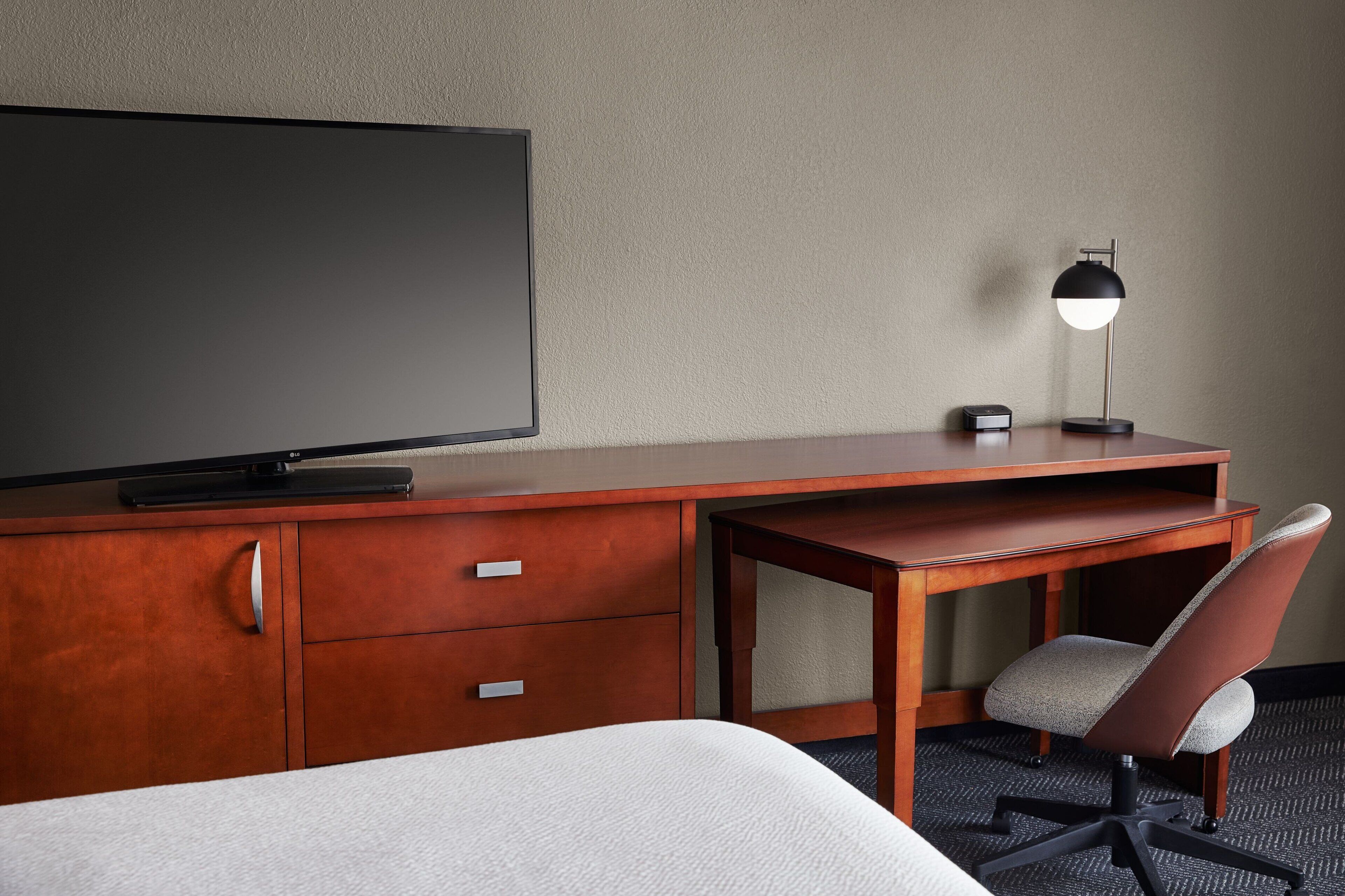 A hotel room with a wooden desk, a television, a desk lamp, a chair, and a bed.