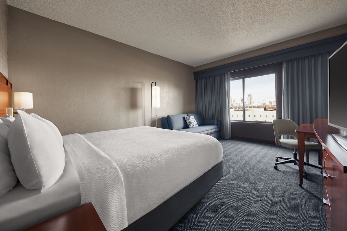 Une chambre d’hôtel avec un grand lit, un bureau, une chaise, une lampe et une vue sur le paysage urbain par la fenêtre.