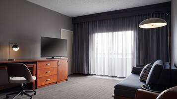 A hotel room with a desk, television, sofa, and large window with curtains.