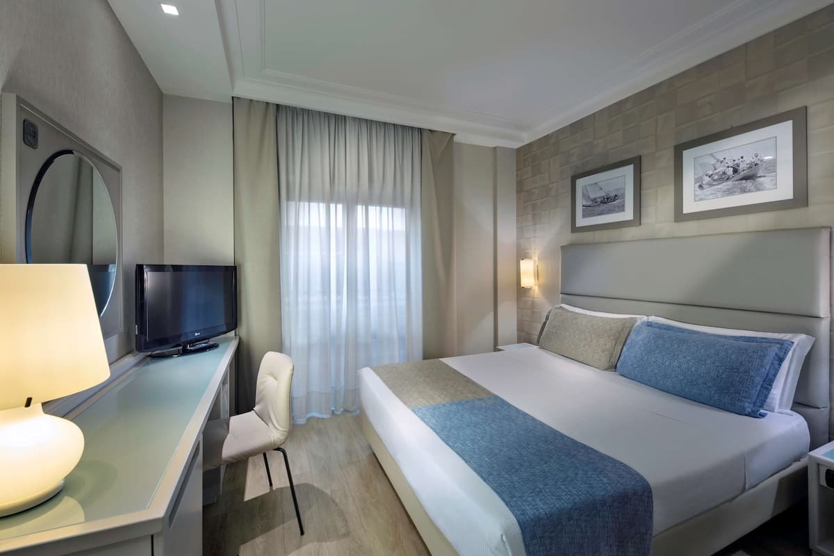 A hotel room with a bed, a desk with a TV, a chair, a window with curtains, and framed pictures on the wall.