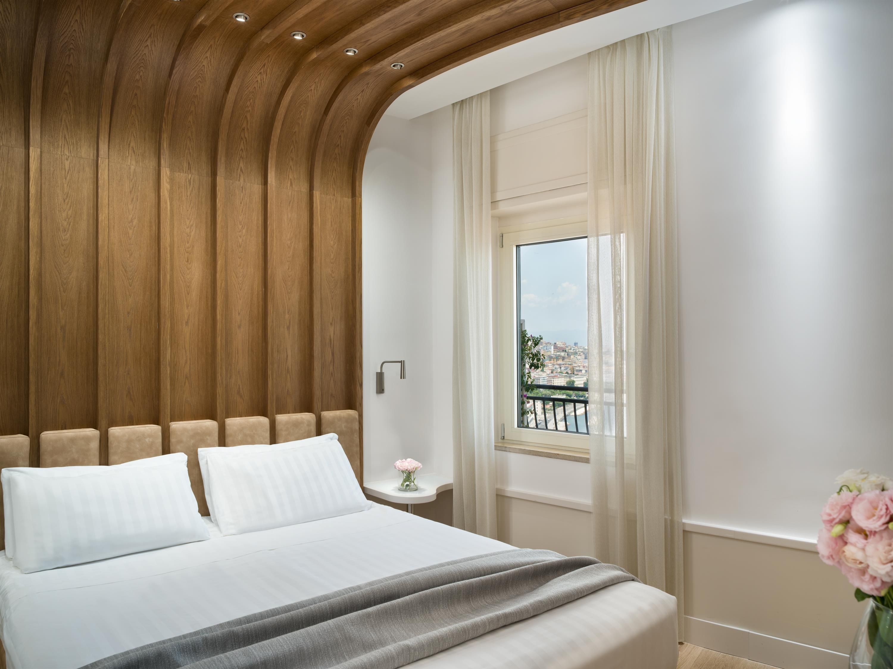 Una camera da letto con testiera in legno, un letto con lenzuola bianche, un comodino con un vaso di fiori e una finestra vista sulla città.