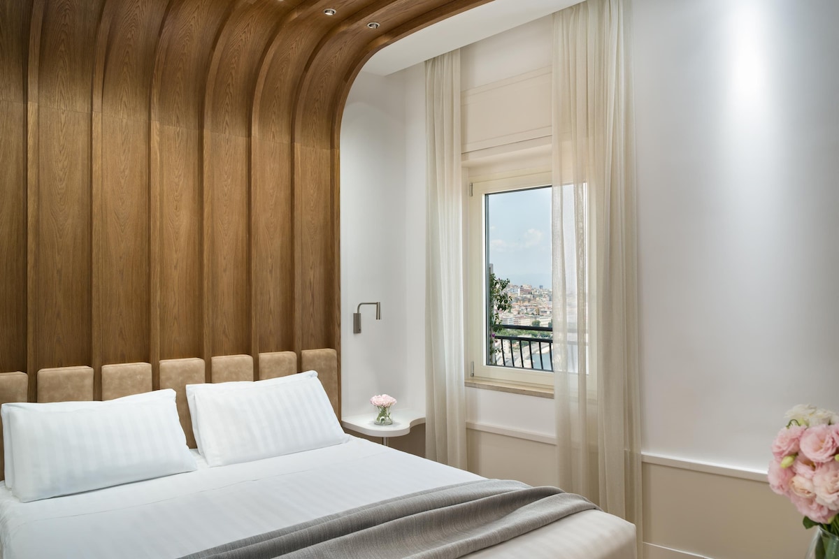 A bedroom with a wooden headboard, a bed with white linens, a nightstand with a vase of flowers, and a window with a view of a cityscape.