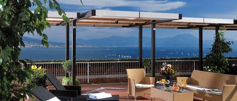A terrace with wicker furniture, a table with drinks, and a view of the sea and mountains.