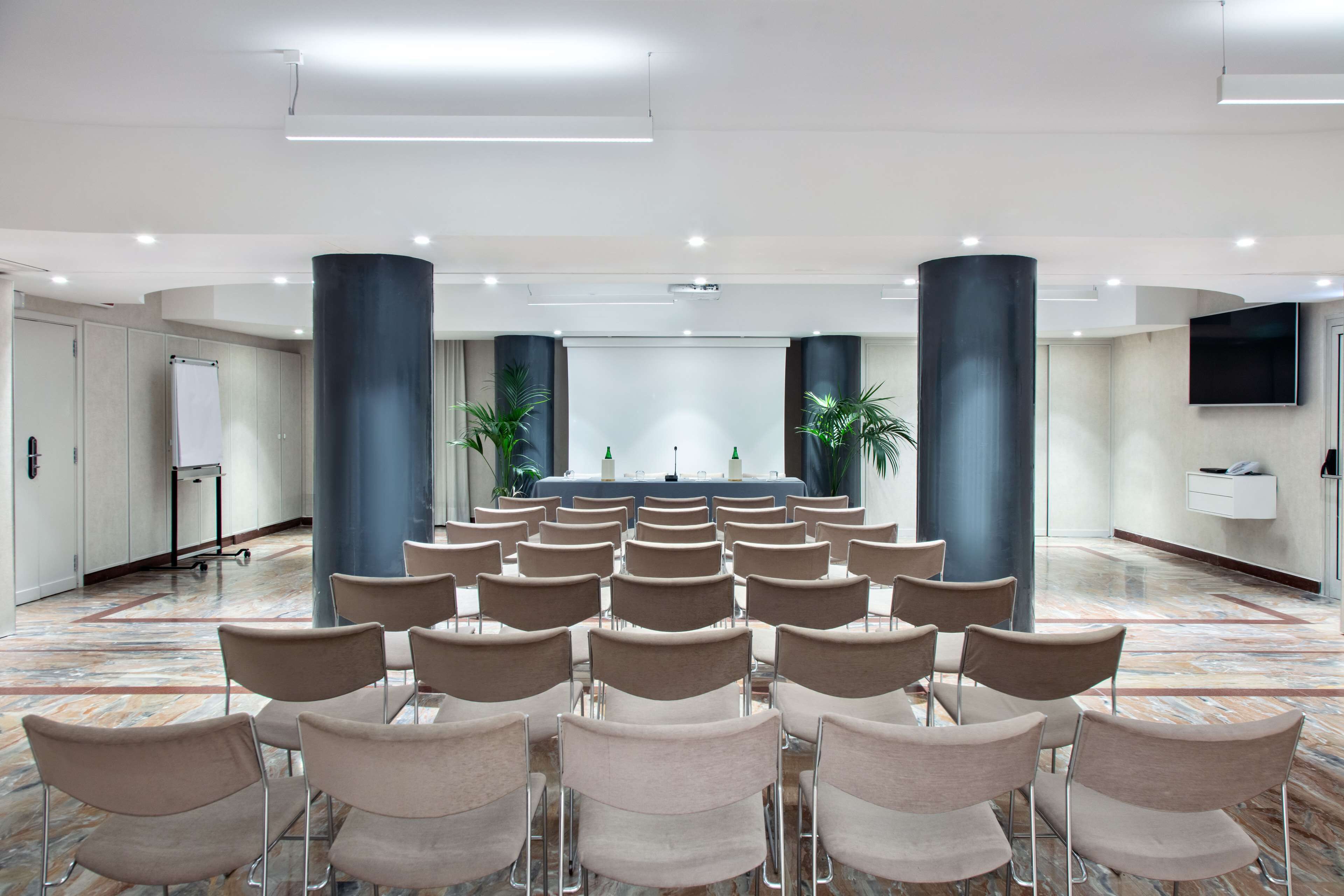 A conference room with rows of chairs facing a stage, large pillars, a whiteboard, and a flat-screen TV on the wall.