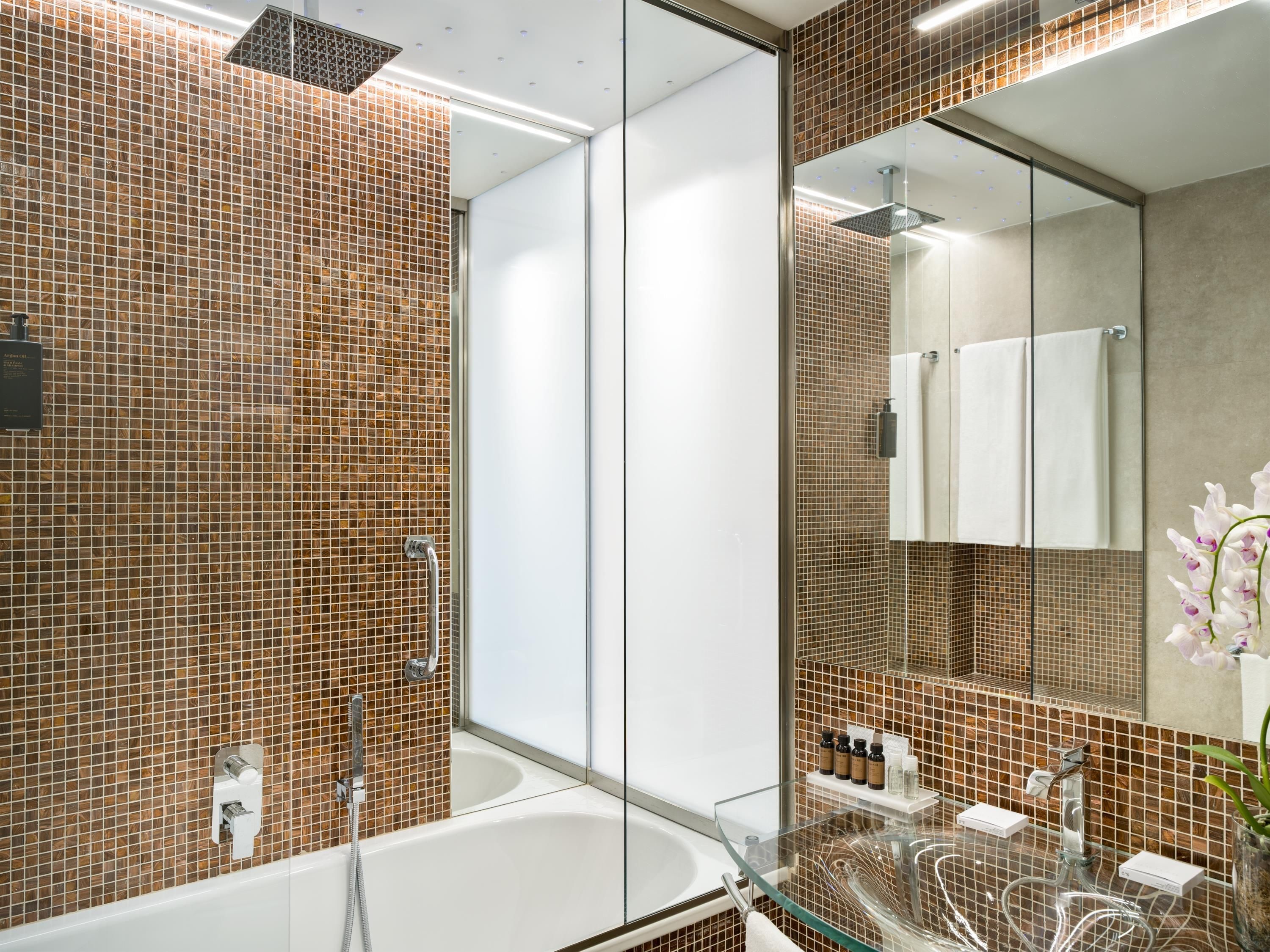 Une salle de bain moderne avec une douche à parois de verre, une baignoire blanche et un meuble-lavabo en verre où sont rangés les articles de toilette.