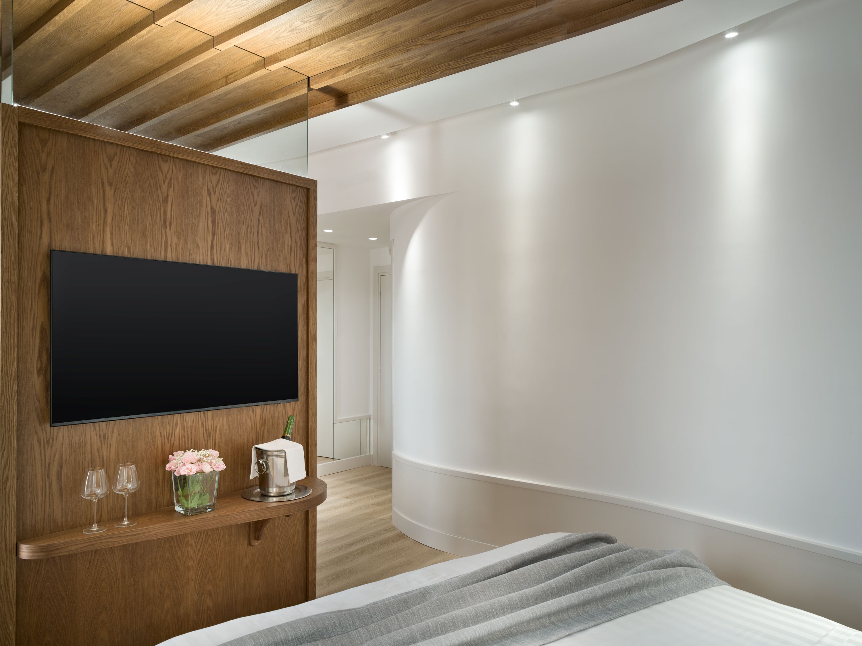 A modern bedroom with a large flat-screen TV mounted on a wooden panel, a bedside table with a vase of flowers, and a bed with a grey blanket.