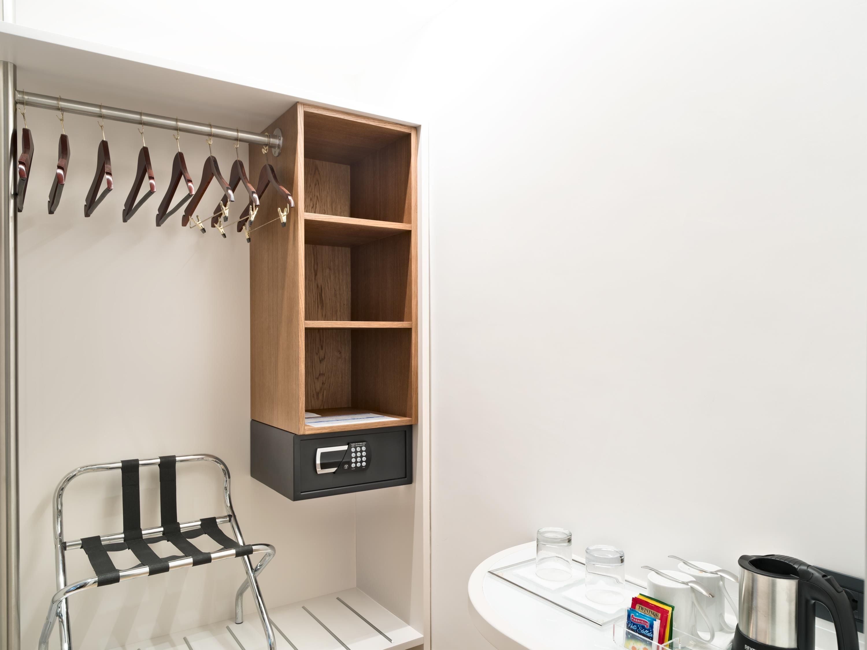 A hotel room with a wooden wardrobe, a metal chair, a safe, and a small table with a kettle and glasses.