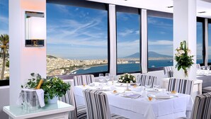A dining area with a table set for two, large windows offering a view of a city and the sea, and a decorative plant on the table. - Hotel Paradiso, BW Signature Collection (Naples)