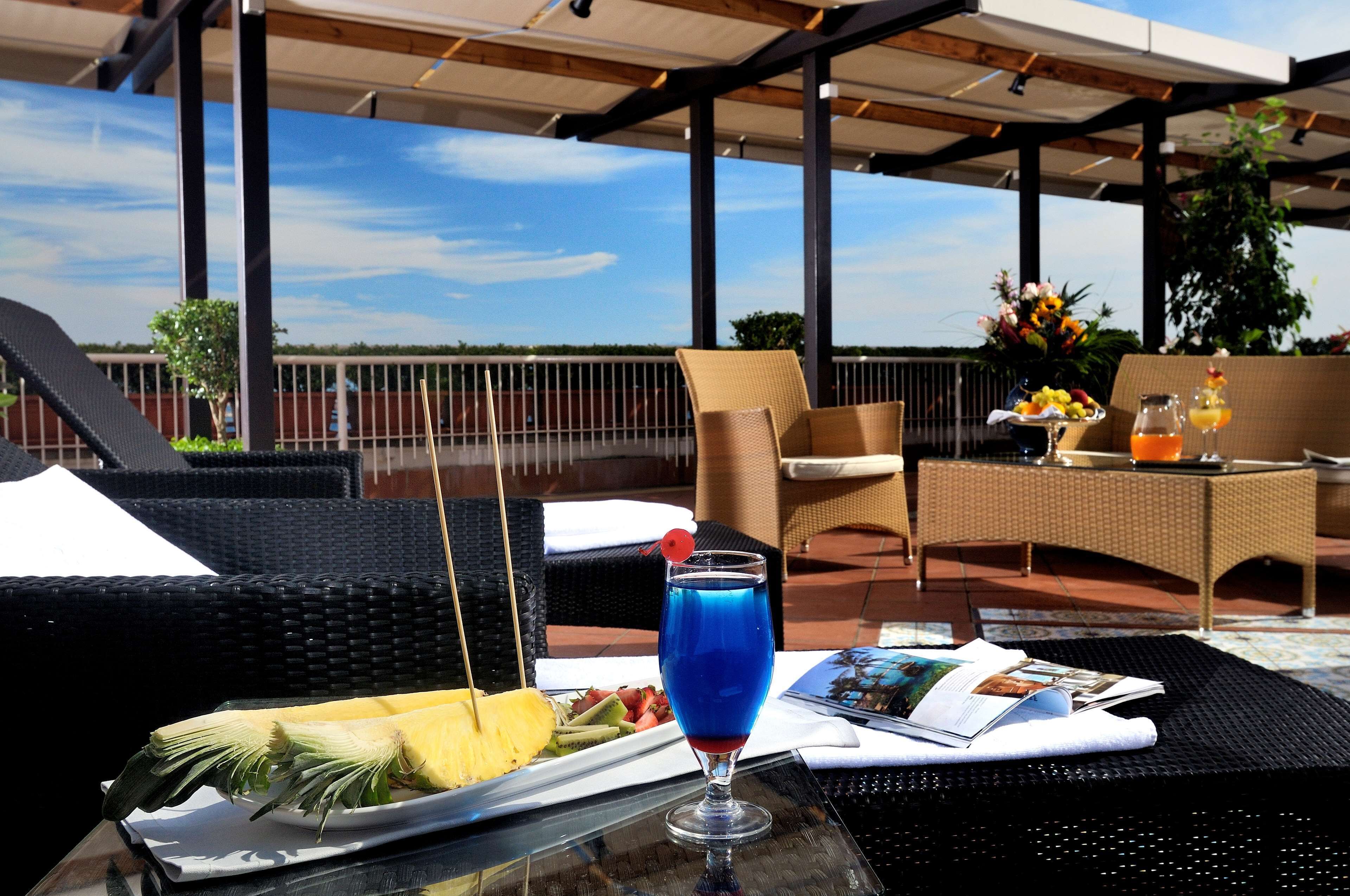 Een tafel met een blauw drankje, een fruitschaal en een tijdschrift op een glazen tafel in een terras met rieten stoelen en uitzicht op de lucht.