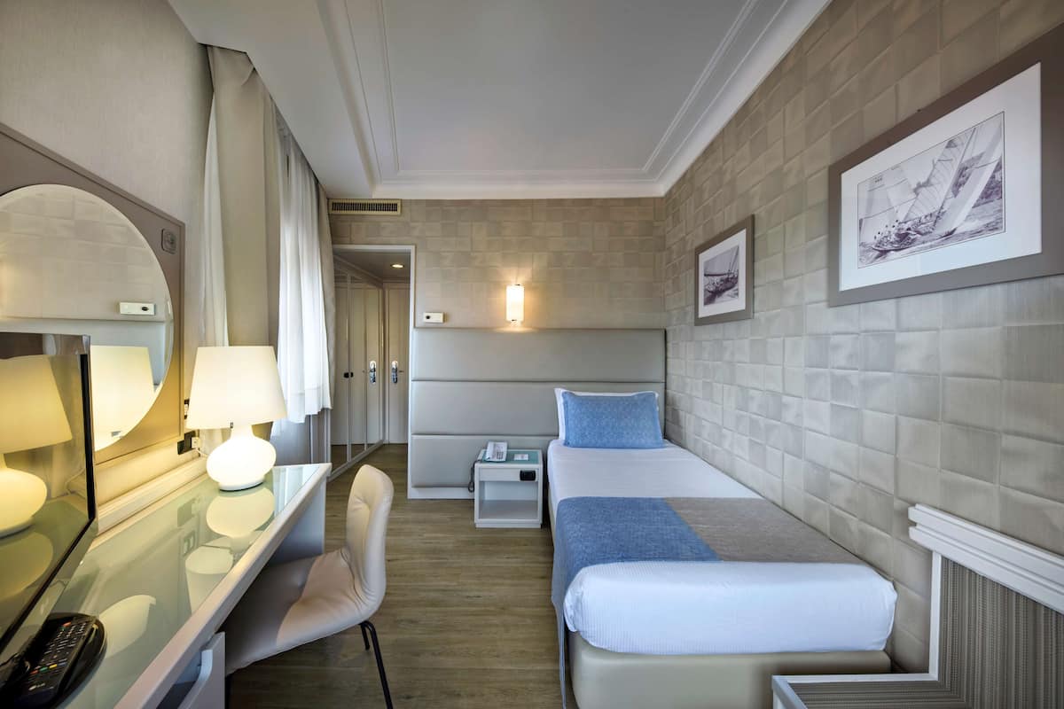 A hotel room with a bed, bedside table, lamp, chair, and a desk with a computer.