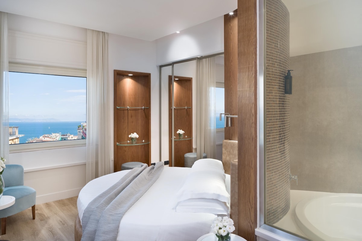 A hotel room with a large bed, a glass shower enclosure, a view of the sea, and a small table with a vase of flowers.