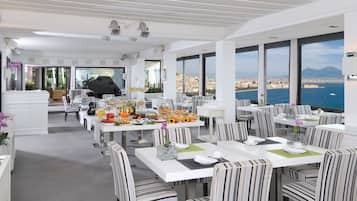 Un'area ristorazione moderna con tavoli bianchi e sedie a righe, un buffet con diversi cibi e ampie finestre con vista sulla città e sul mare.