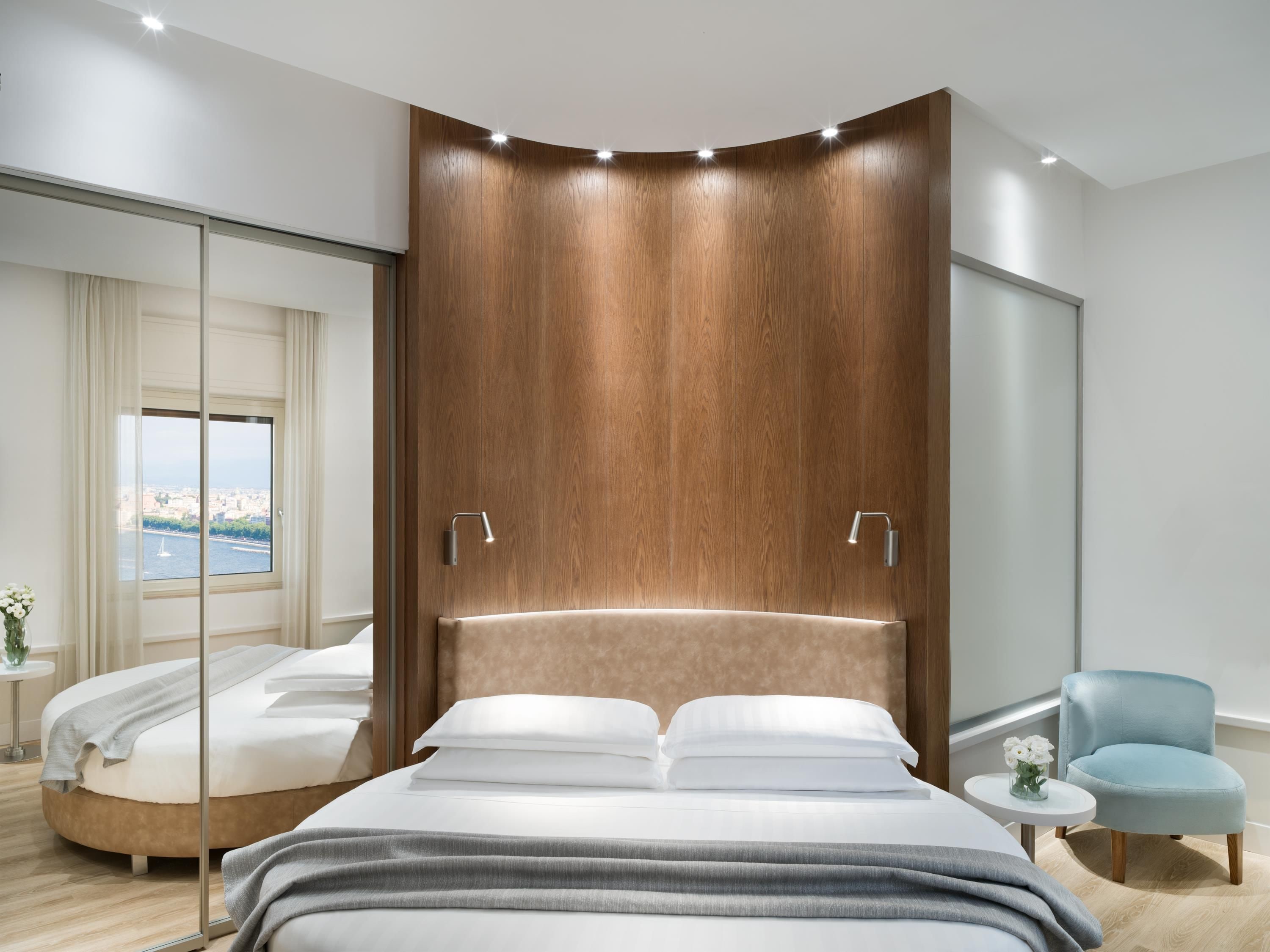 A modern bedroom with a curved wooden headboard, a large bed, a small round table with a vase, and a view of a cityscape through a large window.