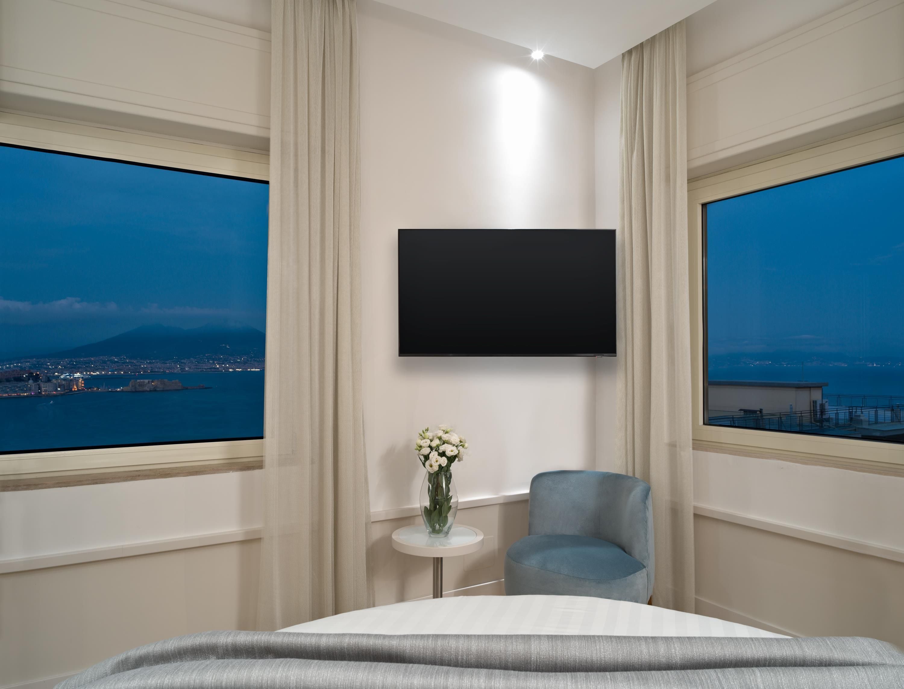 Ein Hotelzimmer mit einem Bett, einem an der Wand befestigten Fernseher, einem kleinen Tisch mit Blumen und großen Fenstern mit Blick auf die Stadt.