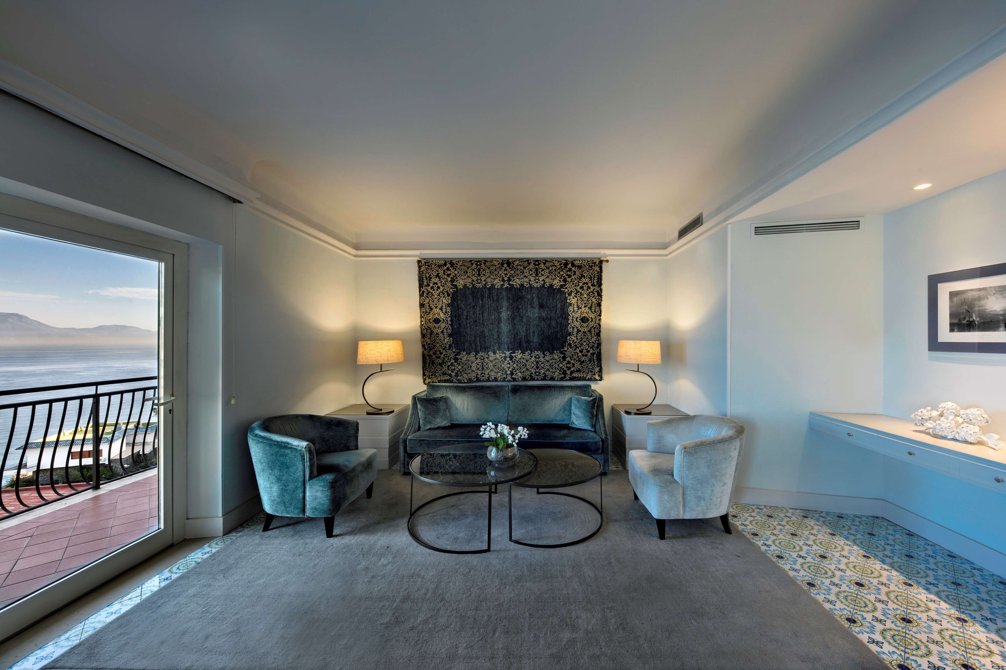 Una habitación de hotel moderna con un ventanal grande que ofrece vista al océano, un sofá mullido, dos sillones, una mesa de centro y un tapete decorativo.