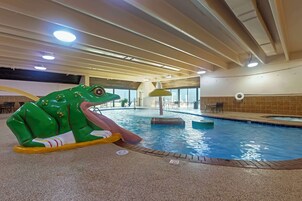 Une piscine intérieure avec un toboggan en forme de grenouille, des luminaires au plafond et une vue sur l’extérieur grâce à de grandes fenêtres.