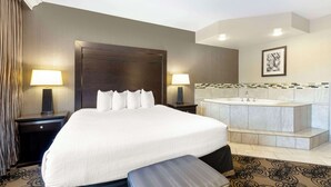 A hotel room with a large bed, two bedside tables with lamps, a small desk, a large window with curtains, a bathroom with a bathtub, and a tiled backsplash.