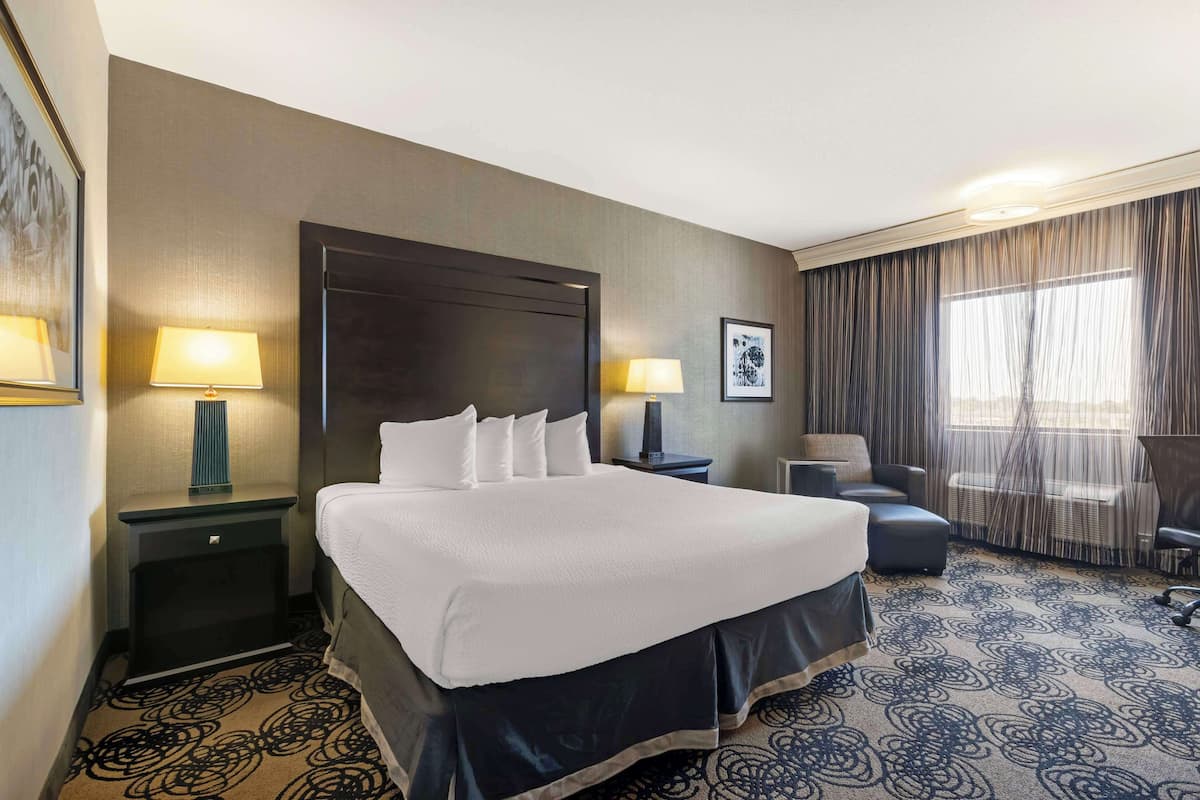 Quarto de hotel com uma cama grande, duas mesas de cabeceira com luminárias, uma cadeira, uma escrivaninha e uma janela grande com cortinas.