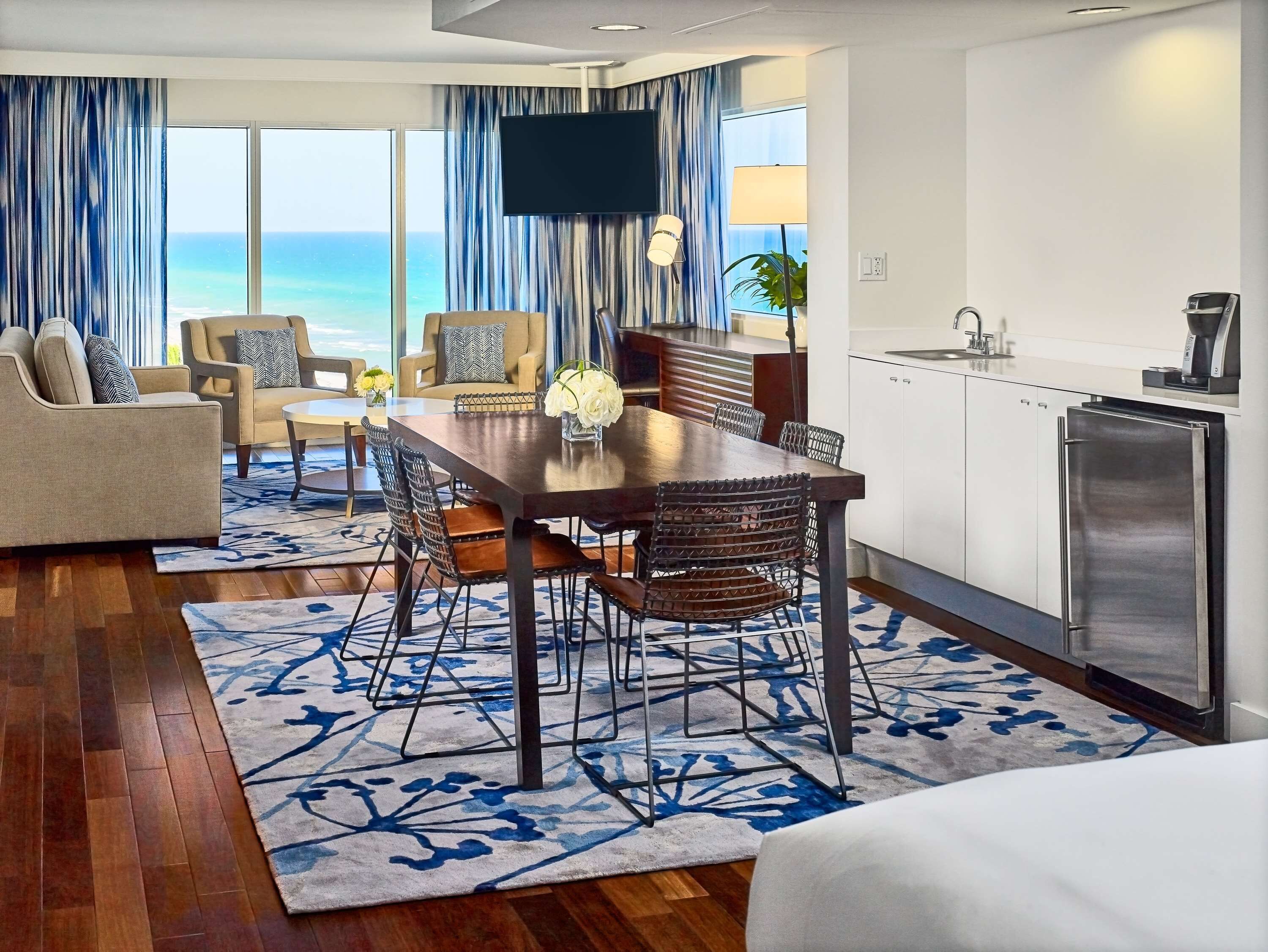 A modern hotel room with a dining area, kitchen, and large windows overlooking the ocean.