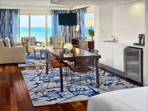 A modern hotel room with a dining area, kitchen, and large windows overlooking the ocean.