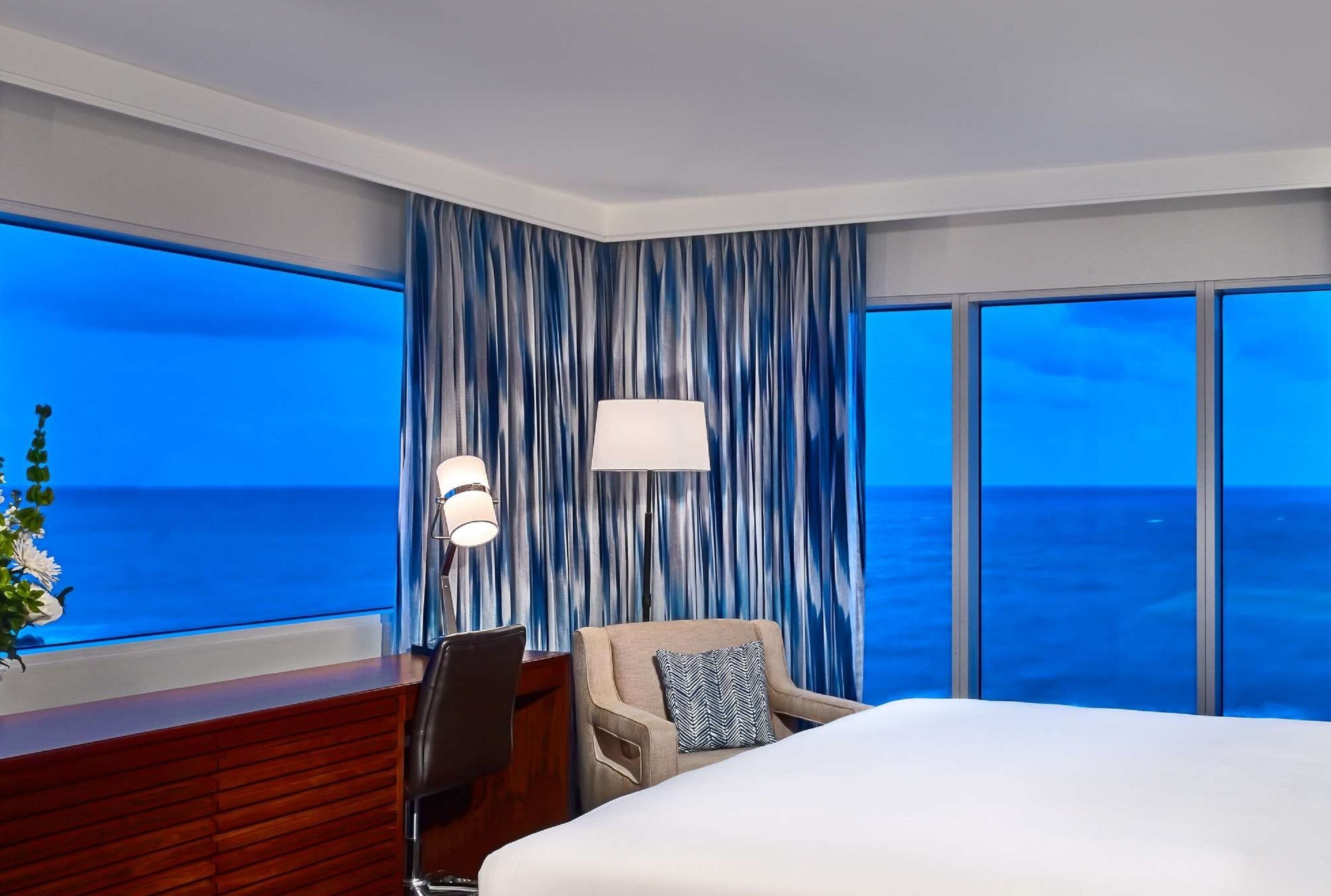 A hotel room with a large bed, a desk with a chair, a lamp, and a view of the ocean.