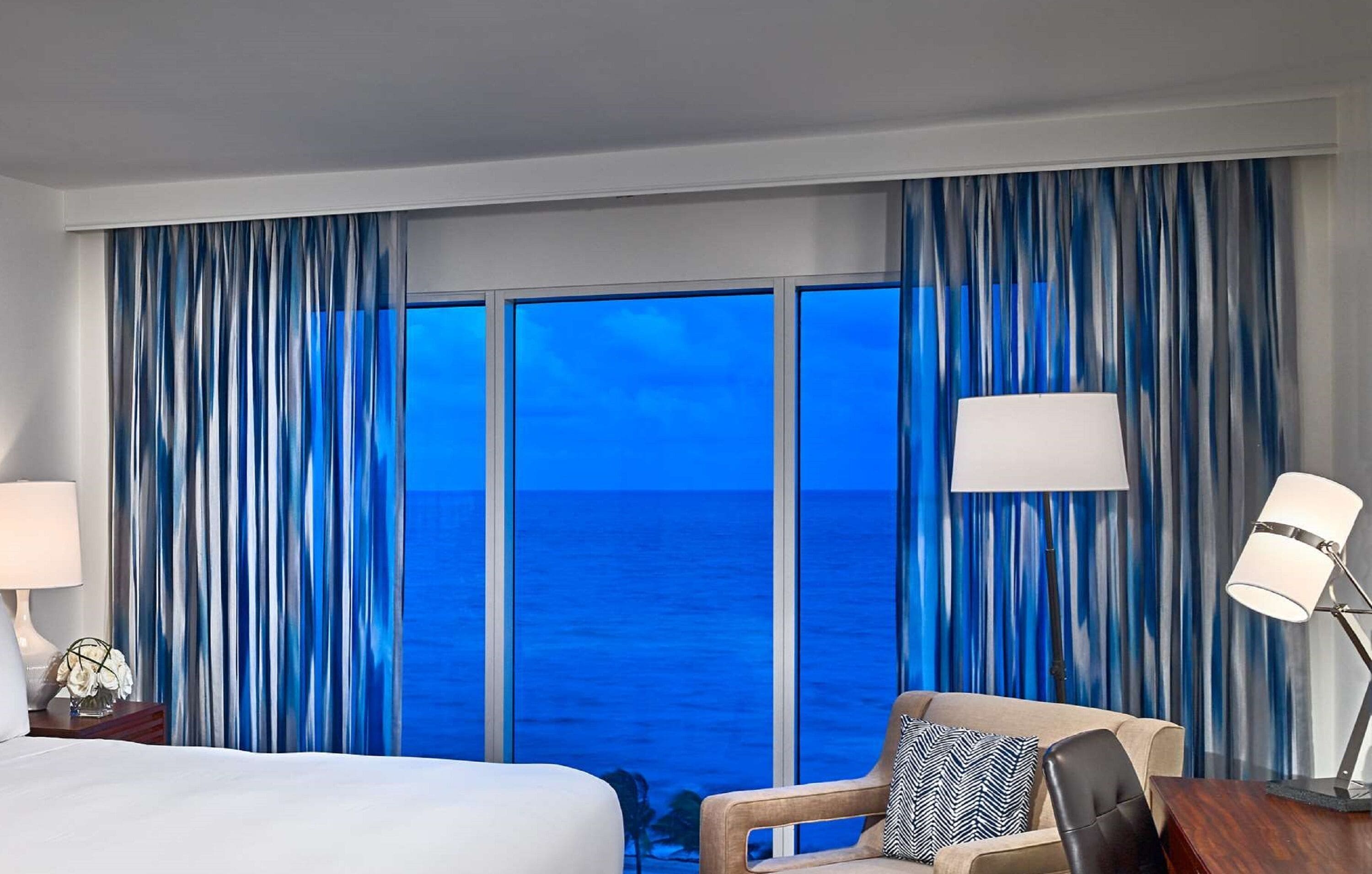 A hotel room with a large bed, two bedside lamps, a chair, and a view of the ocean through floor-to-ceiling windows.