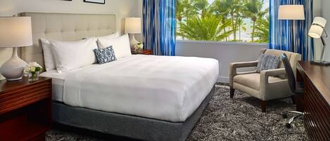 A hotel room with a large bed, two bedside tables with lamps, a chair, a view of palm trees, and two framed artworks on the wall.