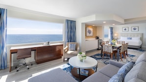 Ett modernt hotellrum med ett stort fönster som vetter mot havet, en mysig sittgrupp med soffa och fåtöljer, ett matbord med stolar och ett litet skrivbord med en stol.