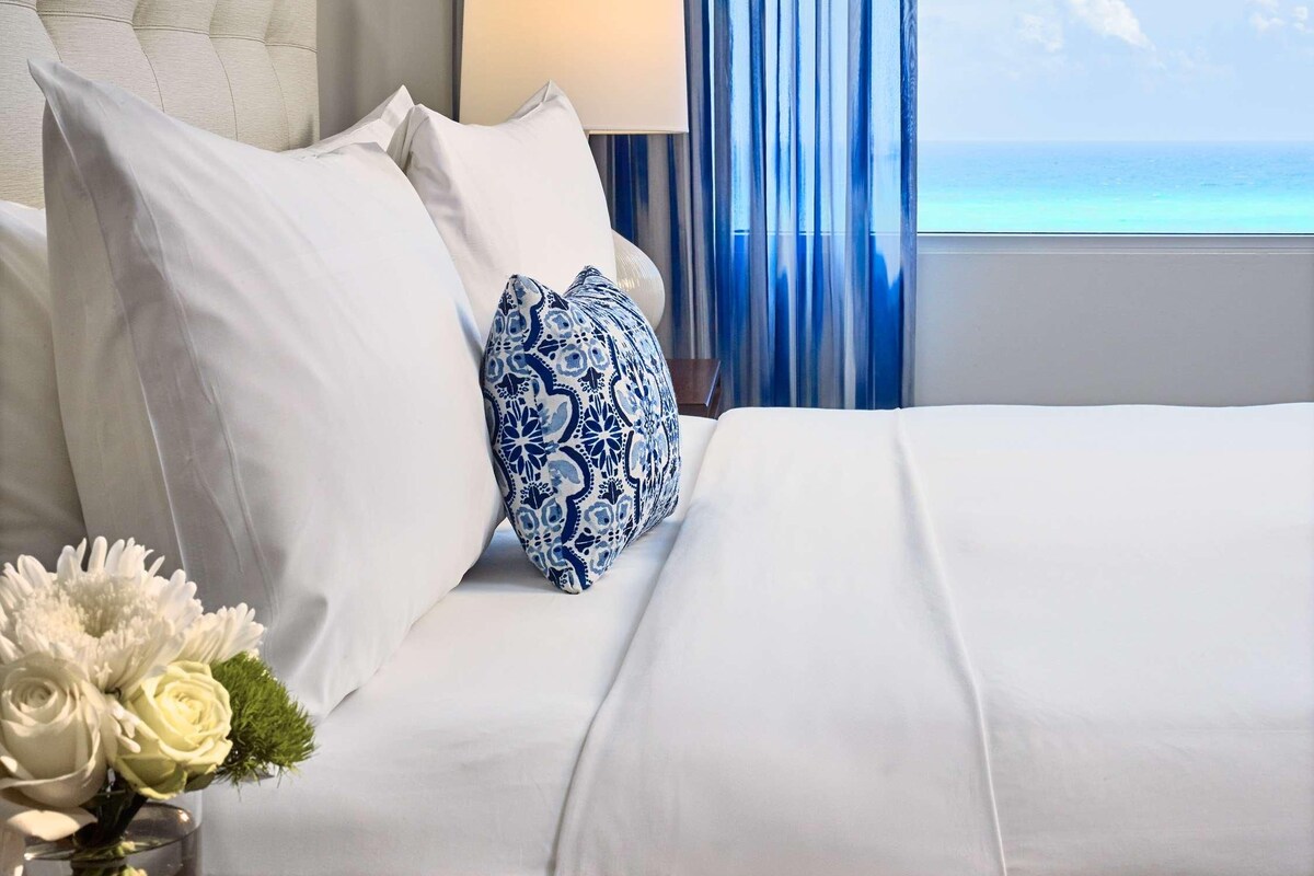 Une chambre d’hôtel avec un lit, une table de chevet, une lampe, une fenêtre avec des rideaux et un tableau encadré au mur.