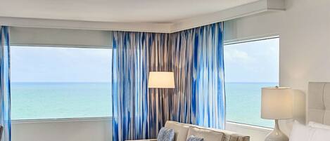 A hotel room with a bed, a sofa, a round table with a vase of flowers, and a view of the sea through large windows.