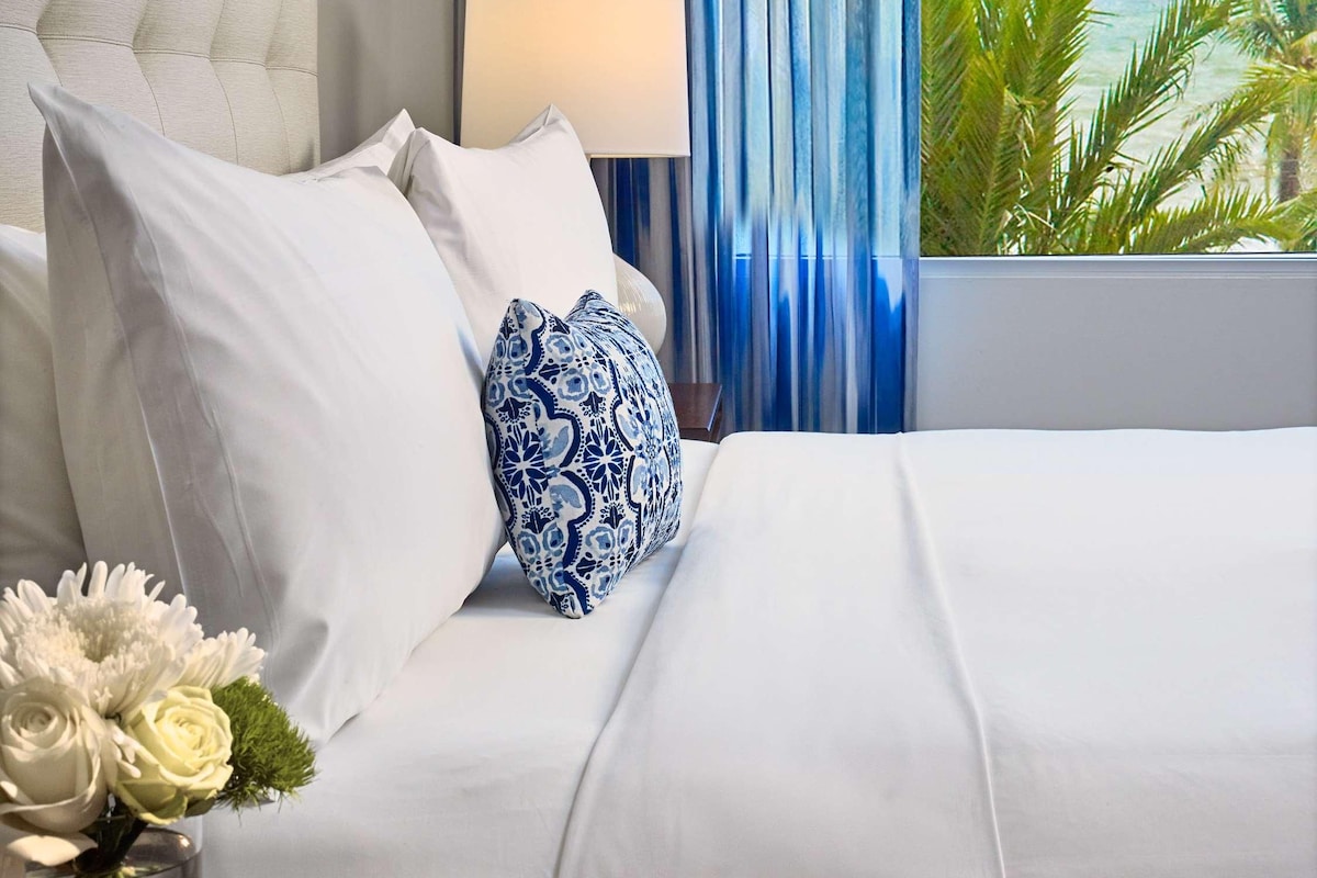 Una camera d'albergo con un letto, un comodino, una lampada, un quadro incorniciato e vista su palme.