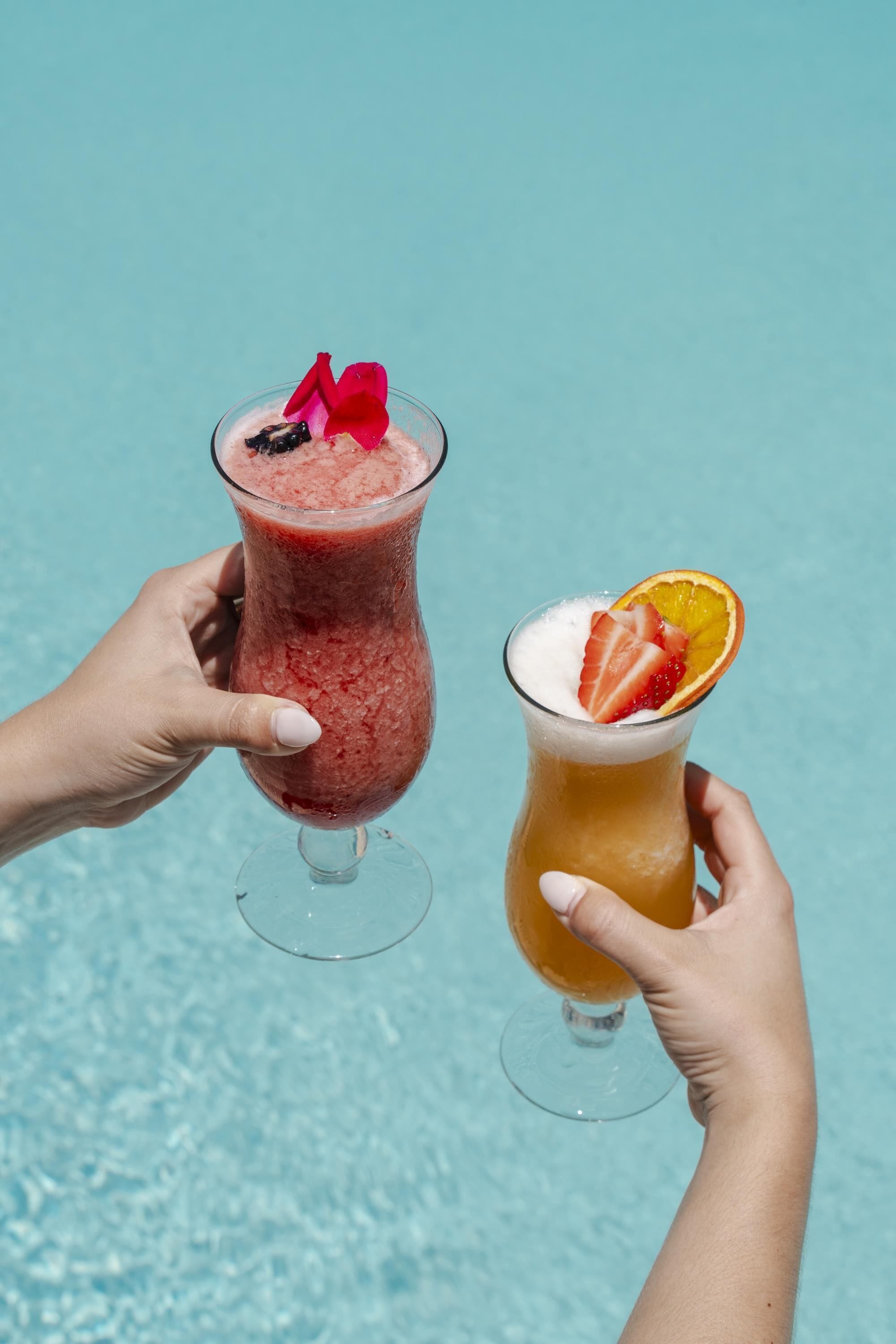 Two hands holding cocktail glasses with fruit garnishes against a blue background.