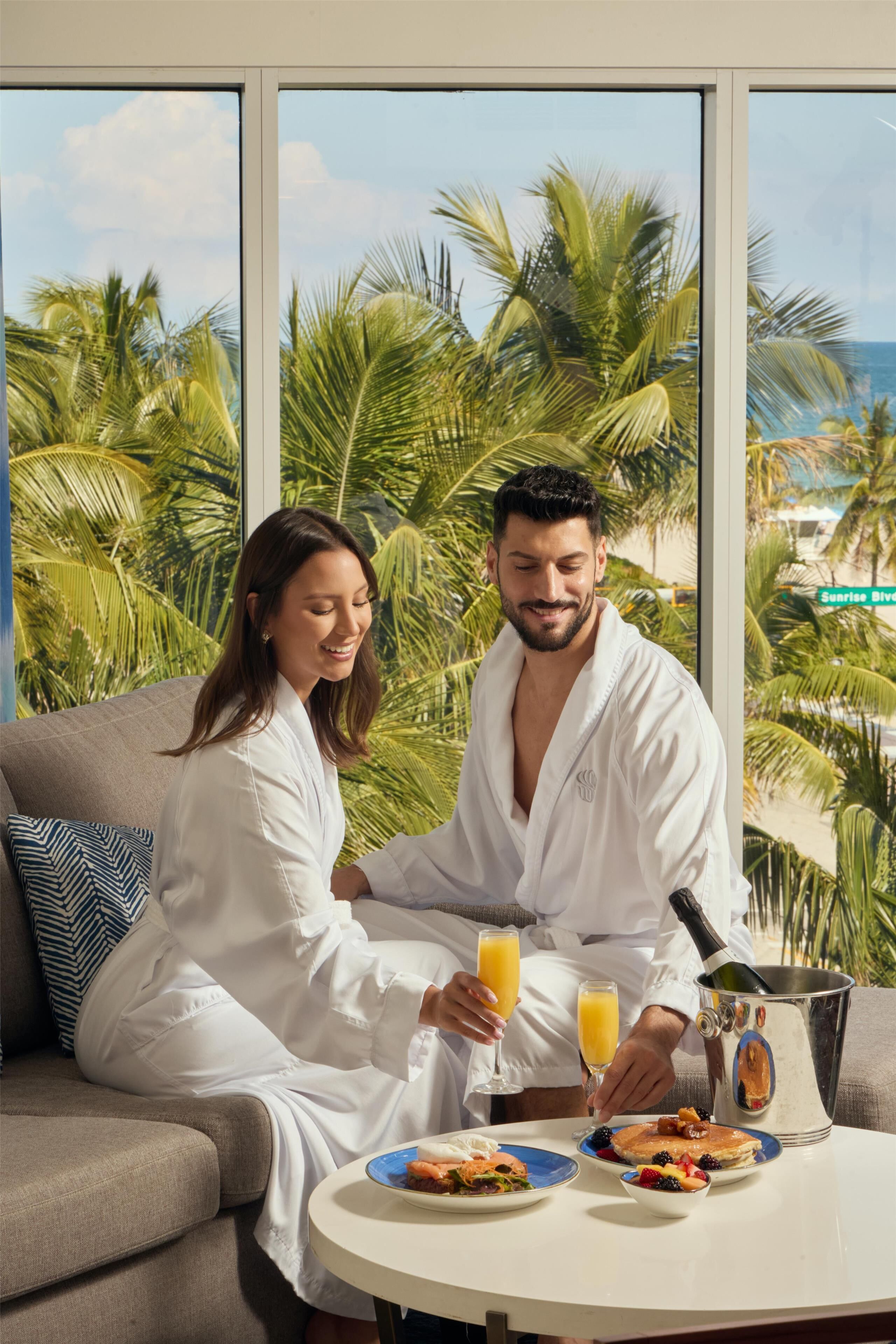 Una pareja con túnicas blancas sentada en un sofá con platos de desayuno, una botella de champán y vasos de zumo de naranja.