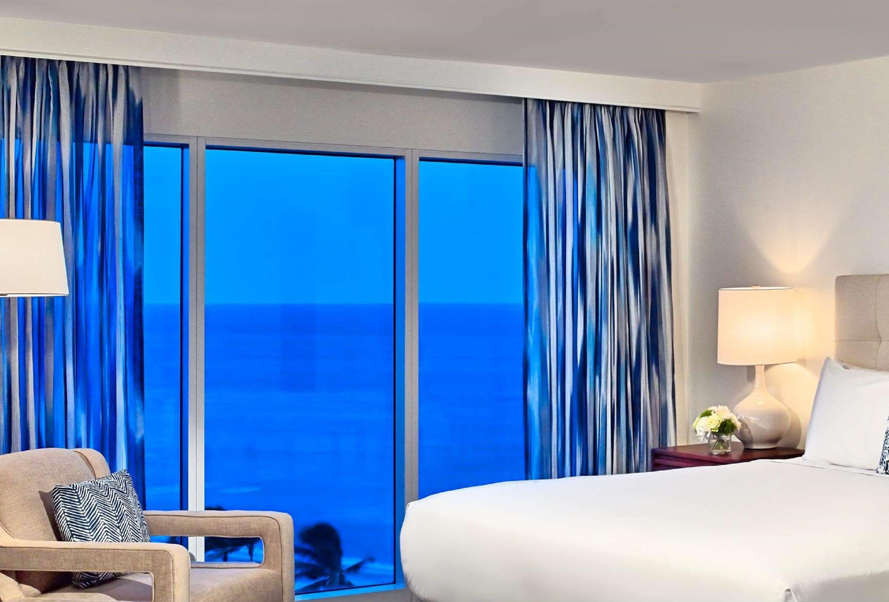A hotel room with a large bed, a chair, a nightstand with a lamp, and a view of the ocean through floor-to-ceiling windows.