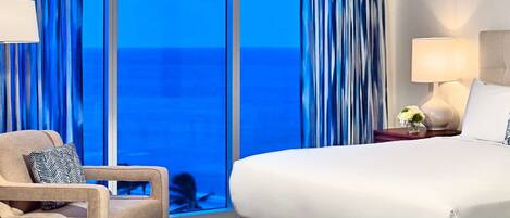A hotel room with a large bed, a chair, a nightstand with a lamp, and a view of the ocean through floor-to-ceiling windows.