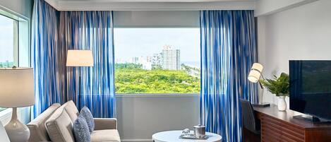 Habitación de hotel con cama, sofá, una pequeña mesa redonda, televisor y vistas a un paisaje urbano a través de amplias ventanas con cortinas azules.