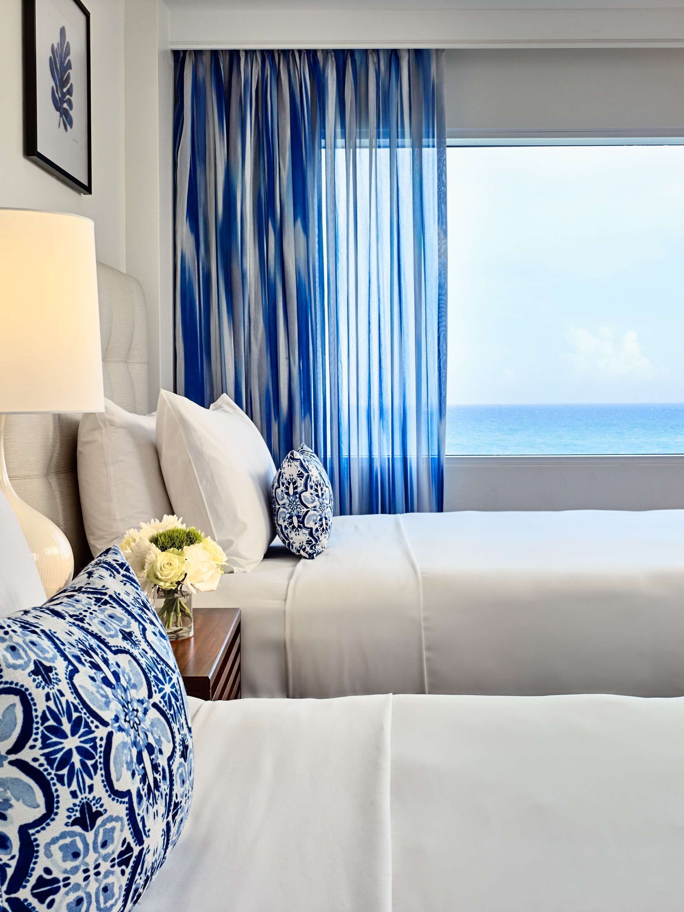 A hotel room with a bed, a chair, a lamp, a vase with flowers, and a view of the ocean.