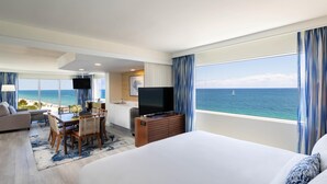 Habitación de hotel con una cama grande, una zona de comedor con mesa y sillas, un televisor y vistas al océano.