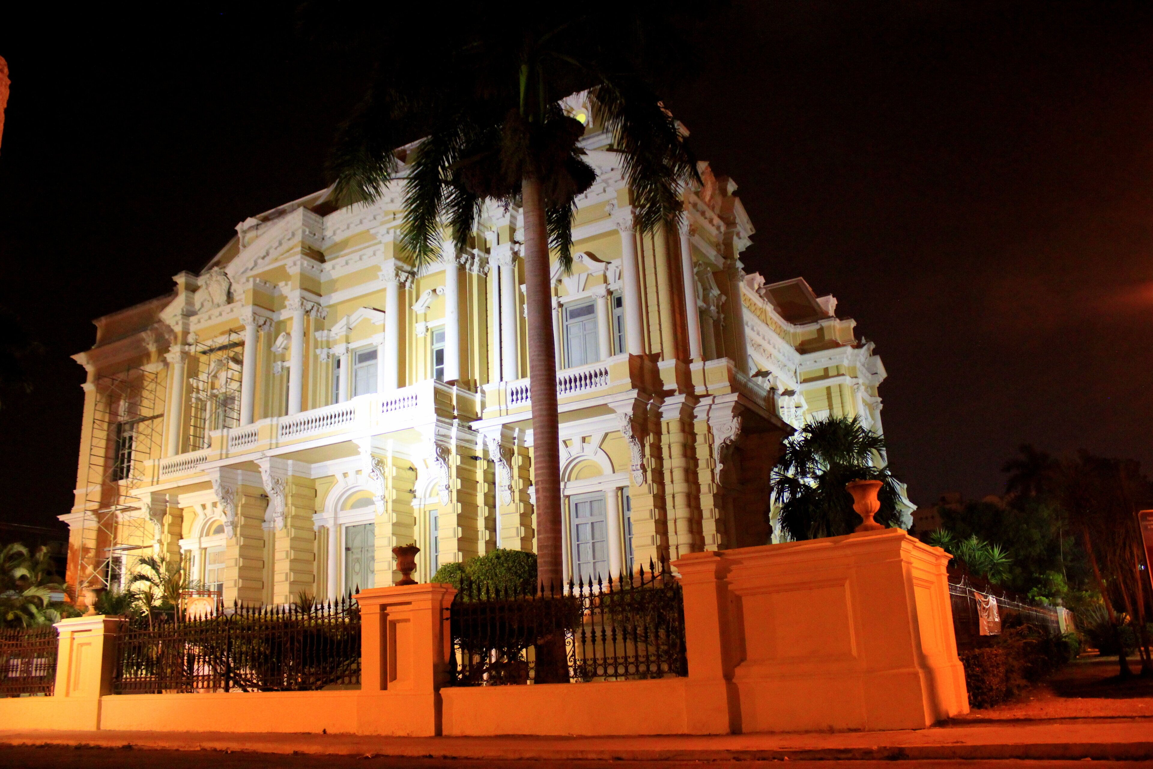A large, ornate building with multiple columns and balconies, surrounded by palm trees and a fence.