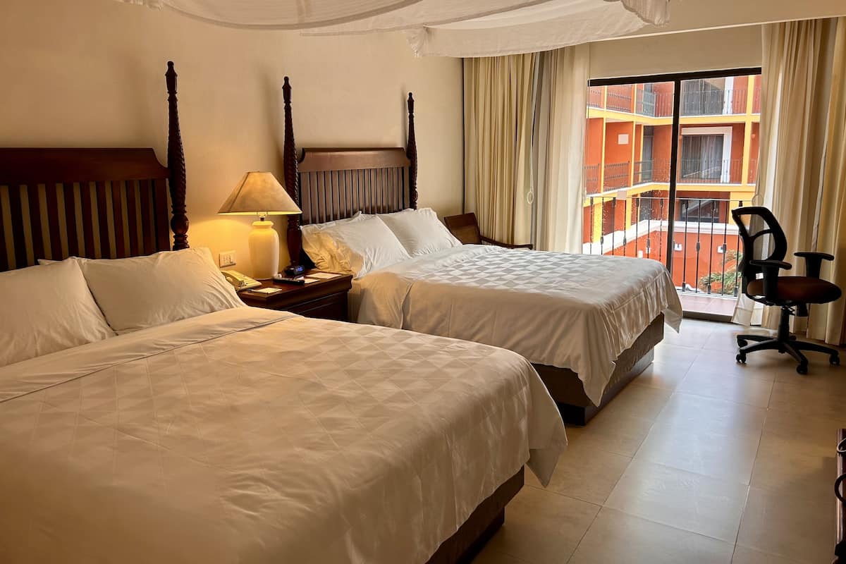 Habitación de hotel con dos camas, una mesita de noche con lámpara, una silla y vista a un edificio con balcones.