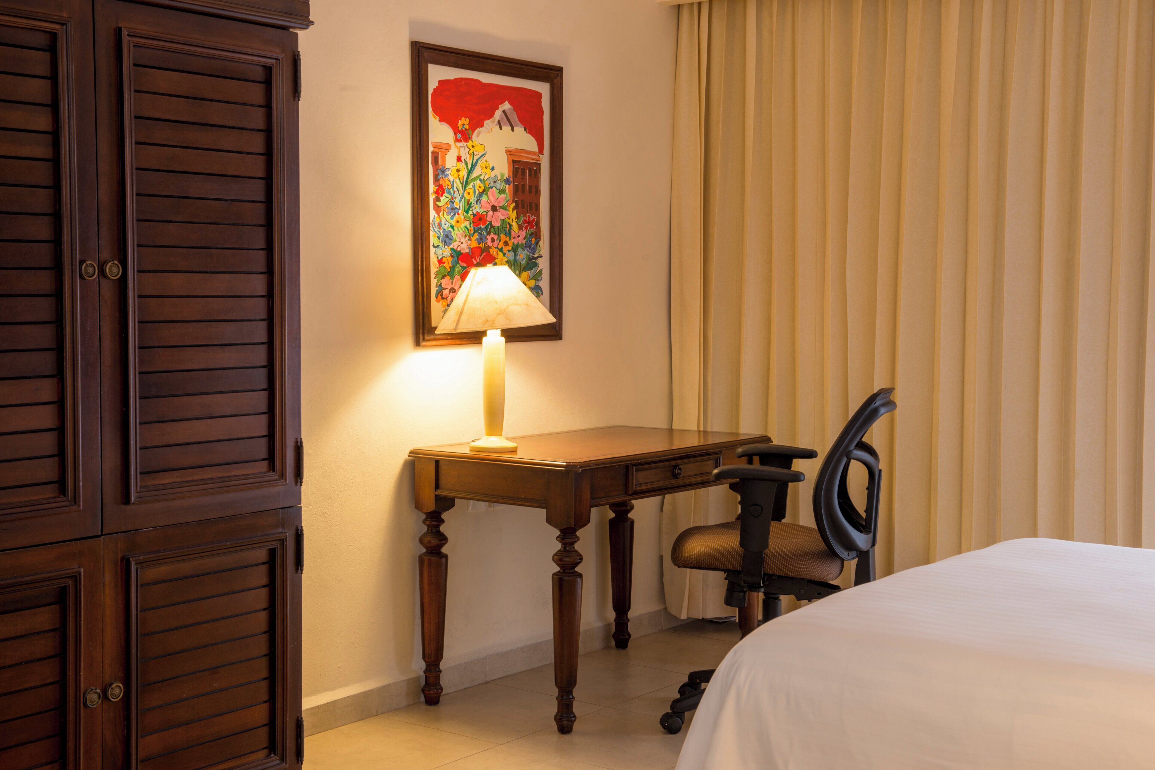 A hotel room with a wooden desk, a chair, a bed, and a painting on the wall.