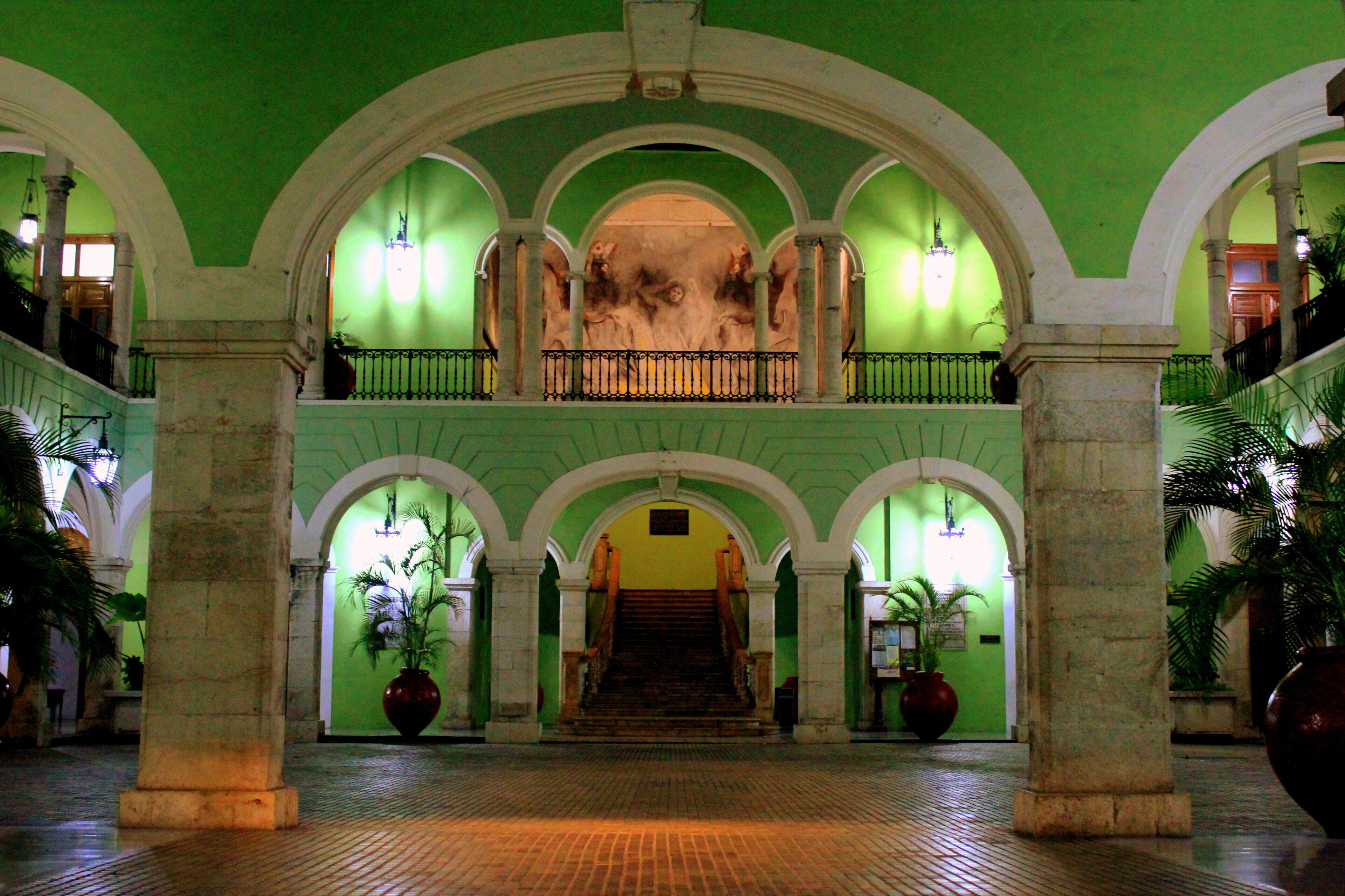 A grand hall with a central staircase, flanked by columns and arches, leading to an upper level.