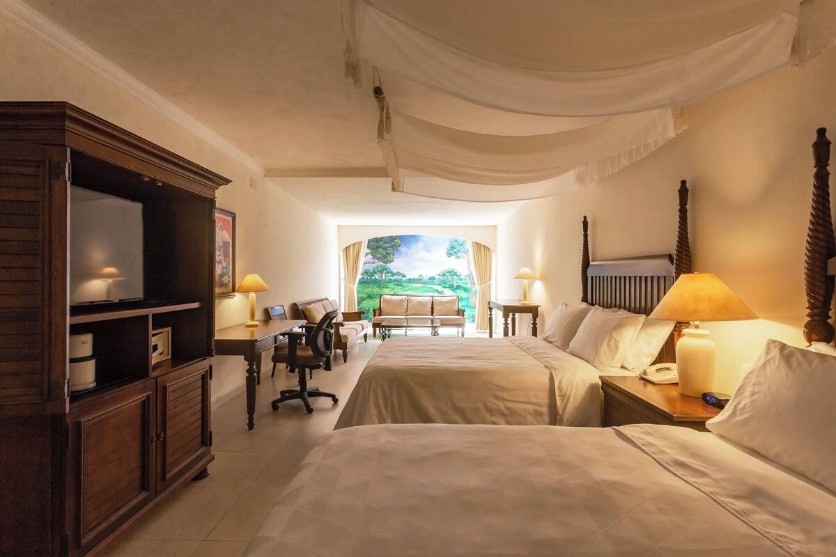 Habitación de hotel con dos camas, un escritorio, una silla, un televisor y vistas a un jardín por la ventana.