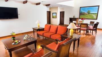 A hotel room with a sofa, armchairs, a dining table, a flat-screen TV mounted on the wall, and a painting above the fireplace.