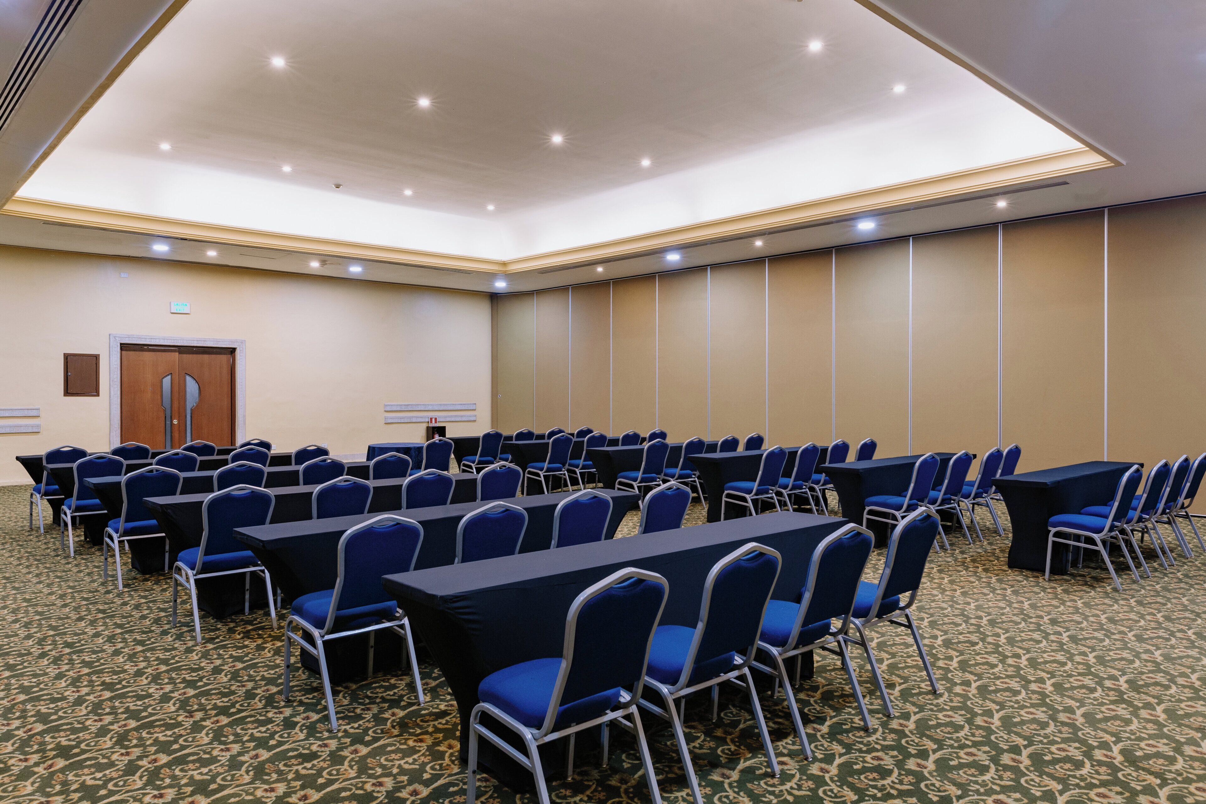 Un salón de conferencias con filas de mesas y sillas, una puerta y suelo alfombrado.