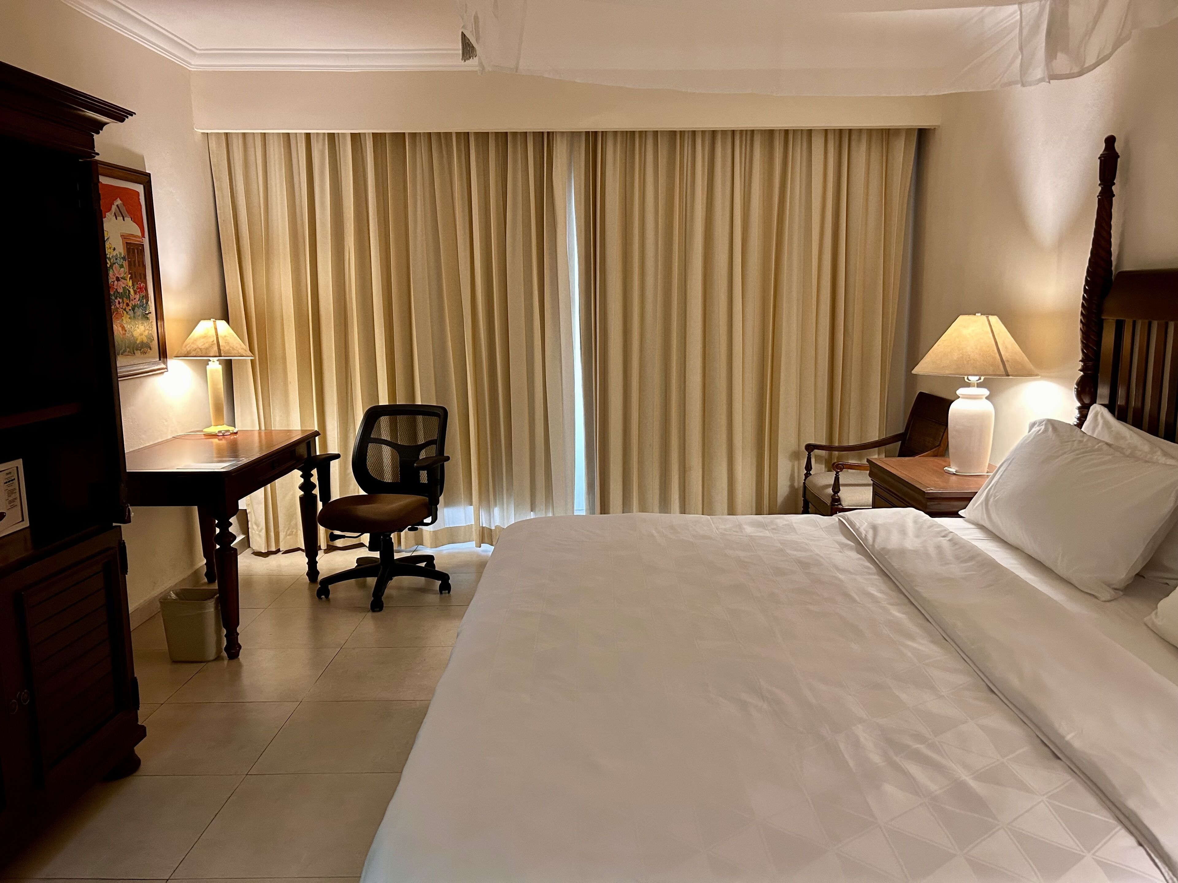 A hotel room with a large bed, a desk, a chair, a nightstand with a lamp, and a window with curtains.