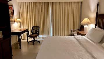 A hotel room with a large bed, a desk, a chair, a nightstand with a lamp, and a window with curtains.